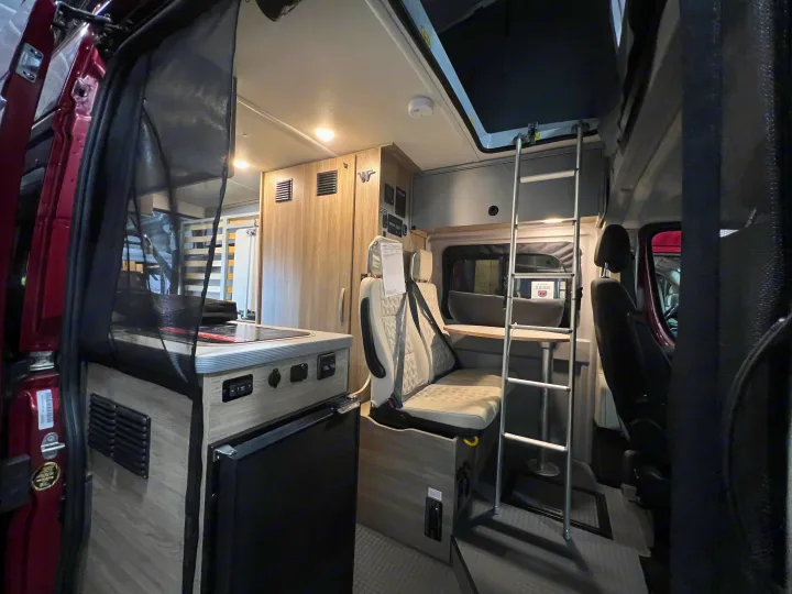 Interior of a modern RV rental, showcasing a compact kitchen, dinette, and ladder leading to a bunk bed for mobile living.. Winnebago Solis 2023