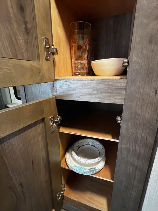 Wood RV rental cabinet open, revealing shelves with glasses, bowls, and paper plates for organized storage.. Cruiser Rv Corp Other 2018