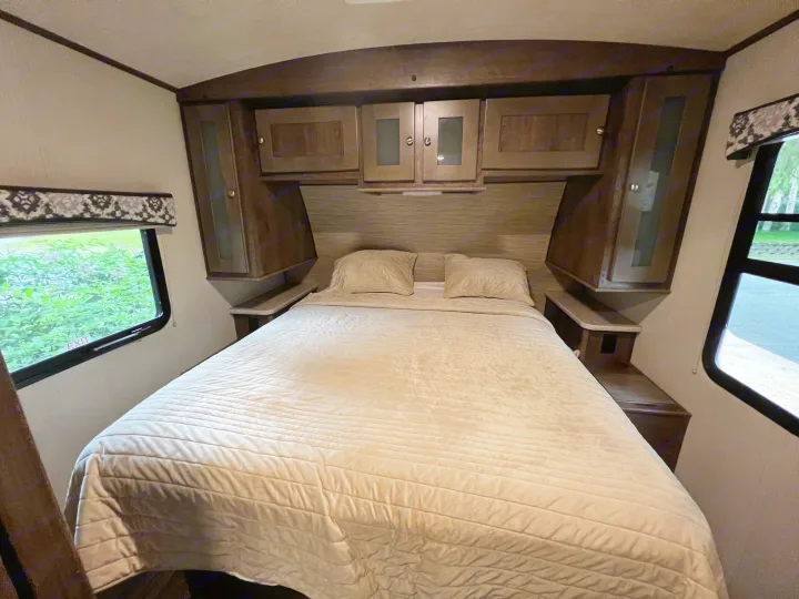 Cozy RV rental bedroom featuring a large bed with light quilted bedding, overhead storage, and windows showing outdoor greenery.. Cruiser Rv Corp Other 2018