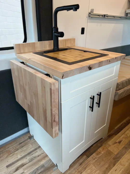 RV rental kitchenette: Light wood counter with modern black faucet, integrated cutting board, and fold-down table. Efficient campervan design.. Ram Promaster 2500 2021