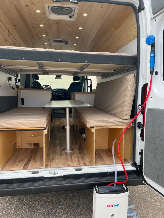 Inside an RV rental: a cozy camper van conversion with a dinette, overhead bed, and a portable HOT TAP shower unit.. Ram Promaster 2500 2021