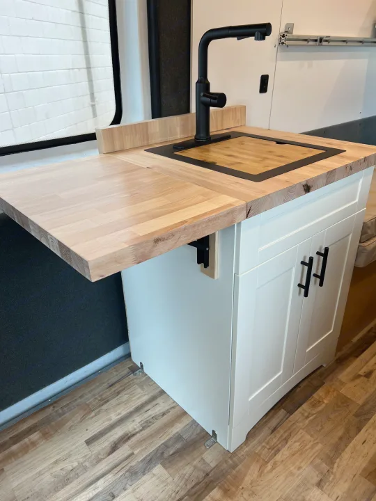 Inside this RV rental: a compact kitchen with a light wood butcher block countertop, modern black faucet, sink, and extendable counter.. Ram Promaster 2500 2021