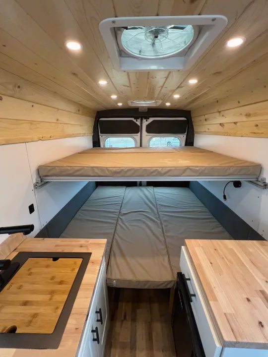 Explore this RV rental's clever interior: stacked beds, wooden ceiling with lights, and kitchenette for comfortable van life.. Ram Promaster 2500 2021