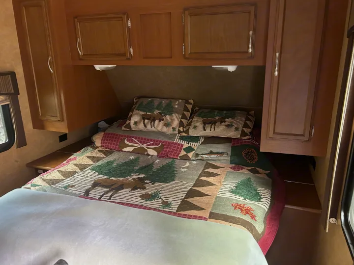 RV rental bedroom with a rustic, wildlife-themed quilt, matching pillows, overhead wooden cabinets, and convenient side shelves.. Starcraft Autumn Ridge 2014