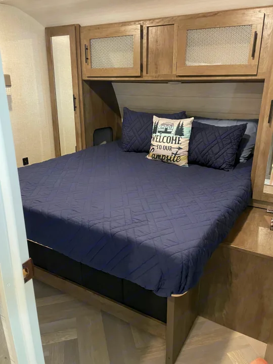 RV rental bedroom features a comfy bed with a "WELCOME TO OUR Campsite" pillow and ample overhead storage.. Forest River Wildwood 2021