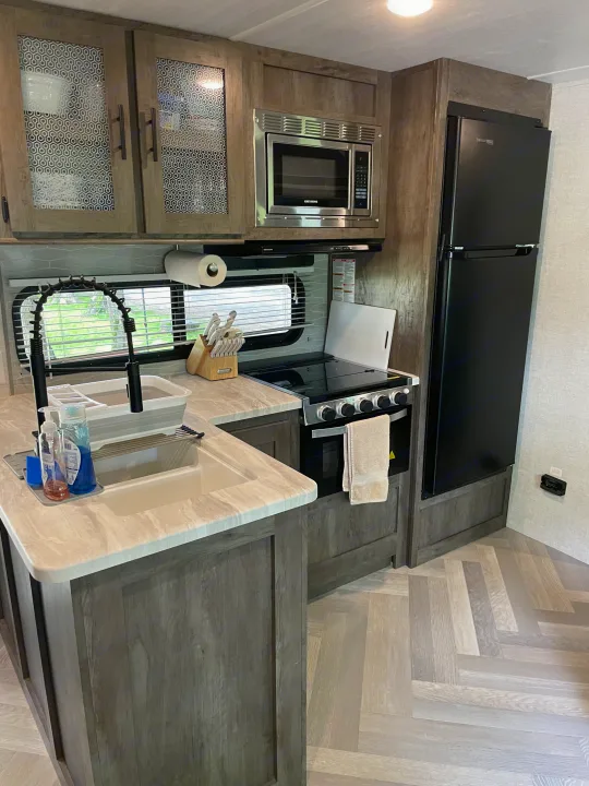 This RV rental kitchen features a modern sink, microwave, stove, and black fridge, complemented by sleek wood-look cabinets and light herringbone flooring.. Forest River Wildwood 2021