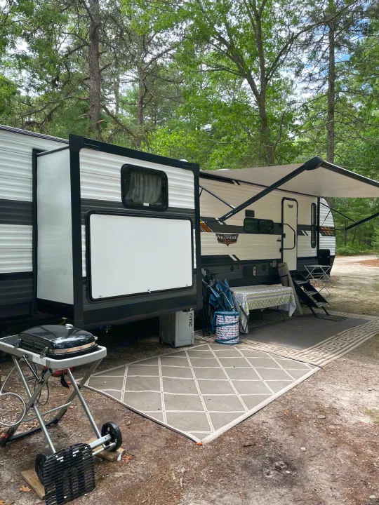 Enjoy a cozy rv rental setup at a shaded campsite, complete with an extended awning, rugs, and grill.. Forest River Wildwood 2021