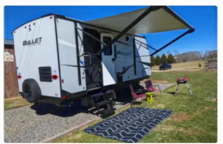 RV rental. White travel trailer with awning, steps, outdoor mat, chairs, and grill at a sunny campsite.. Keystone RV Bullet Crossfire 2023