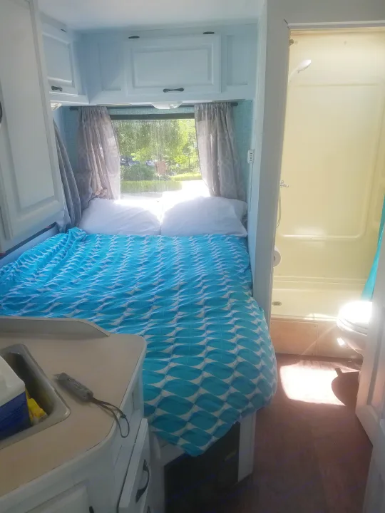 Inside a bright rv rental: a cozy bed with blue bedding, overhead cabinets, a sink, and a compact bathroom with shower and toilet.. four winds majestic 2004