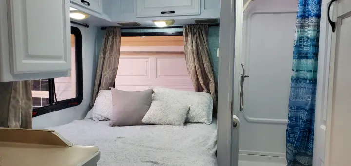 Cozy RV rental interior with a bed, patterned curtains, white cabinets, and a compact bathroom with a blue shower curtain.. four winds majestic 2004