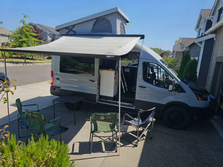 Gray RV rental campervan with pop-top roof and extended awning, set up with camping chairs and table for outdoor comfort.. Grit Overland GO A/T 2022