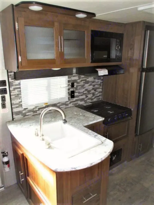Inside an RV rental, a modern kitchen with a sink, microwave, gas stove, wood-look cabinets, and decorative tile backsplash.. Gulf Stream Kingsport 2020