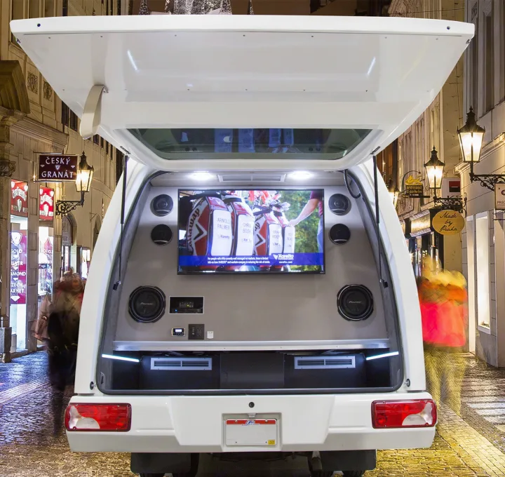 An RV rental's custom tailgate opens to a mobile entertainment system with a large TV and speakers, illuminated at night on a European street.. Custom Coach Mauck2 2015
