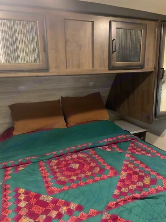 Inside this RV rental's cozy bedroom: a green & pink patterned quilt, brown pillows, and overhead storage cabinets.. Heartland RVs Mallard 2021
