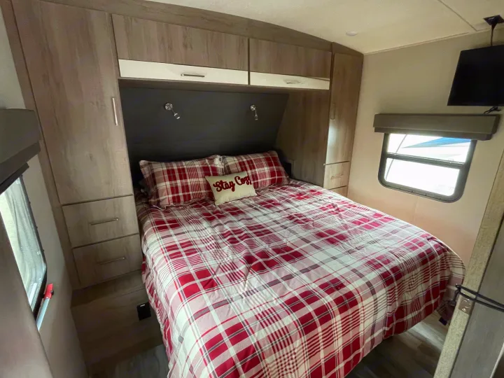 This RV rental bedroom features a plaid bed, ample built-in storage, a TV, and a window, offering a cozy retreat.. Grand Design Imagine 2022