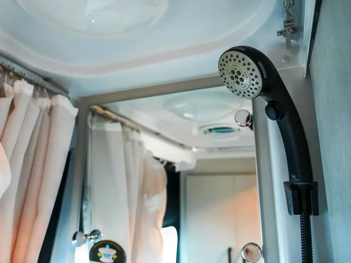 Adjustable black showerhead in a compact RV rental bathroom, with light privacy curtains and reflective surfaces.. Thor Dazzle 2024