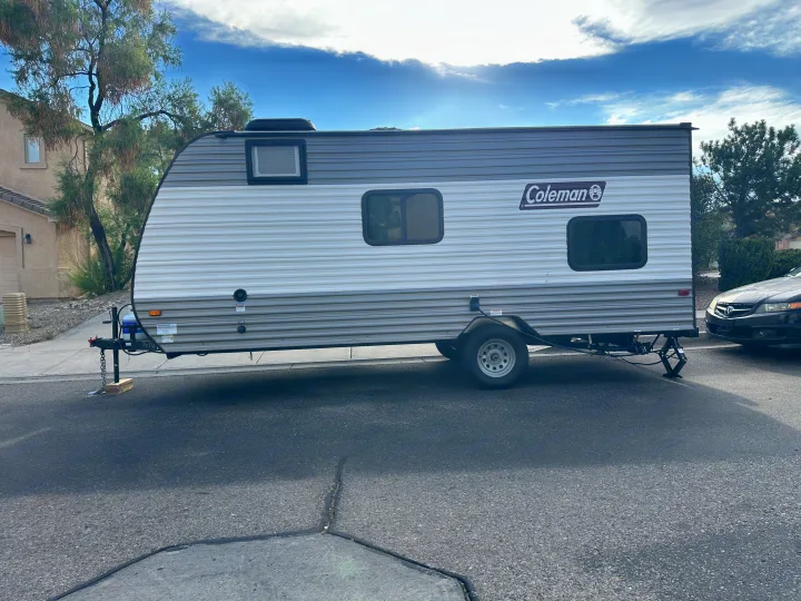 A gray and white Coleman travel trailer (RV rental) is parked on a residential street, supported by jacks under a sunny sky.. Coleman Lantern 17B 2024