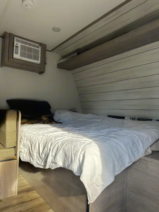 RV rental bedroom interior: a white-covered bed, wall-mounted AC, and wood paneling offer a comfortable stay.. Coleman Lantern 17B 2024