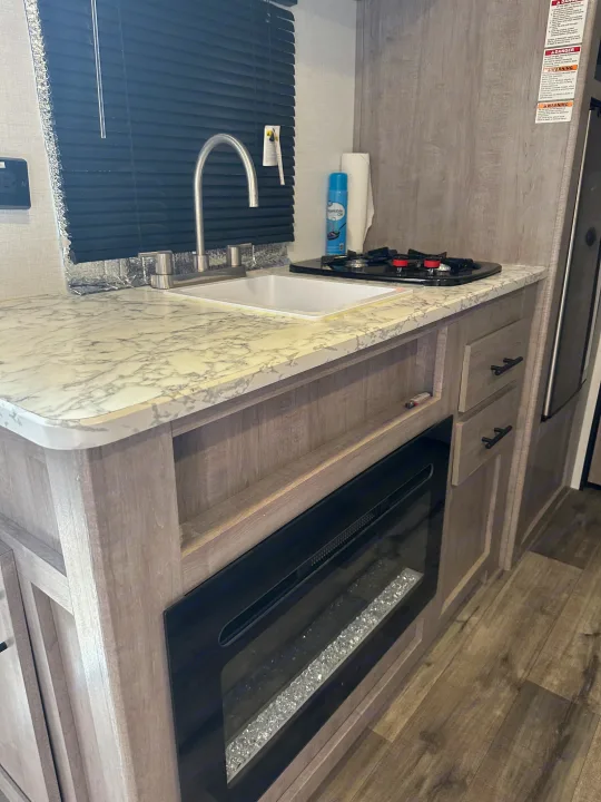 Modern rv rental kitchen with a sink, stovetop, and electric fireplace. Features wood-look cabinetry and marbled countertops.. Coleman Lantern 17B 2024