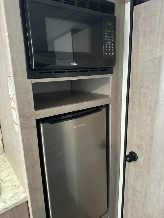 This rv rental features a built-in black microwave above an open shelf and a stainless steel mini-fridge in wood-grain cabinetry.. Coleman Lantern 17B 2024
