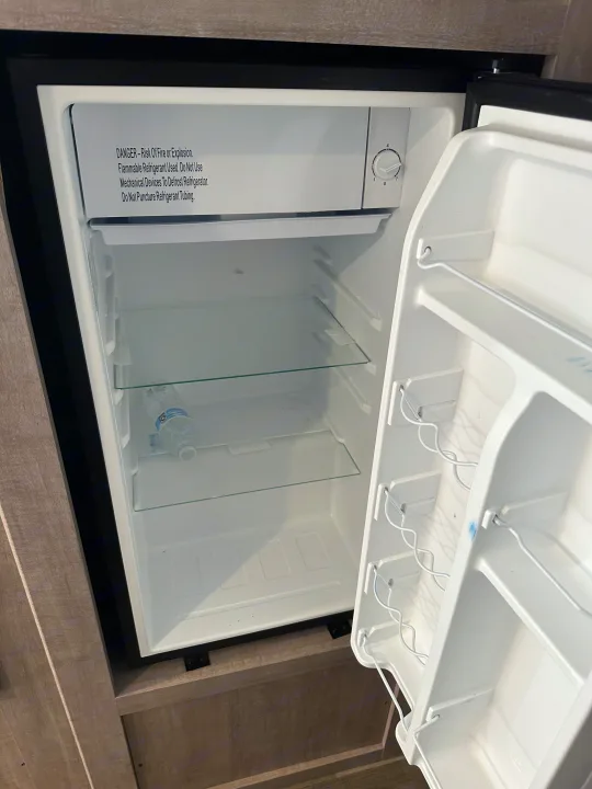 Open rv rental mini-fridge with warning label, temperature dial, and empty shelves. A water bottle is inside.. Coleman Lantern 17B 2024