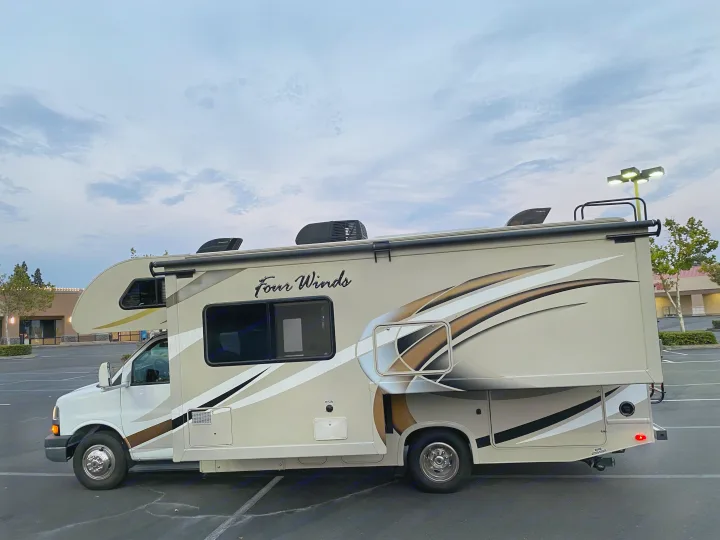 A beige Class C Four Winds RV rental with brown and white stripes, parked in a vast lot under a cloudy sky. Explore travel!. Thor Four Winds 2017