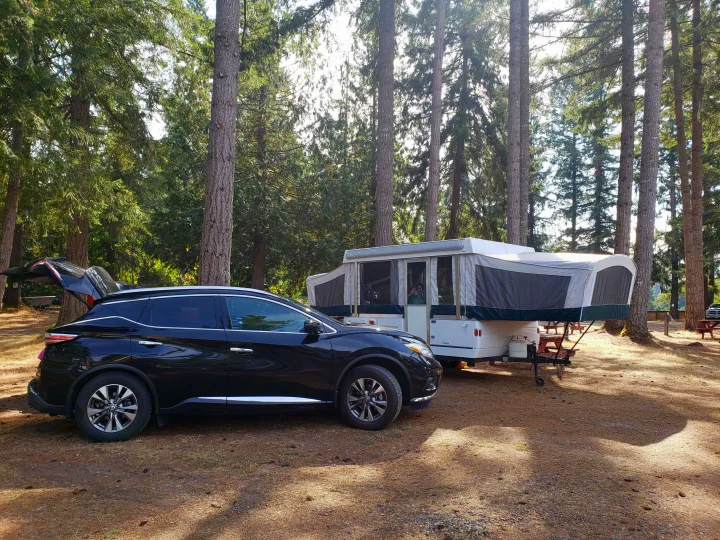A black SUV and pop-up RV rental are set up for a sunny day camping in a lush forest campsite.. Coleman Sedona 1999