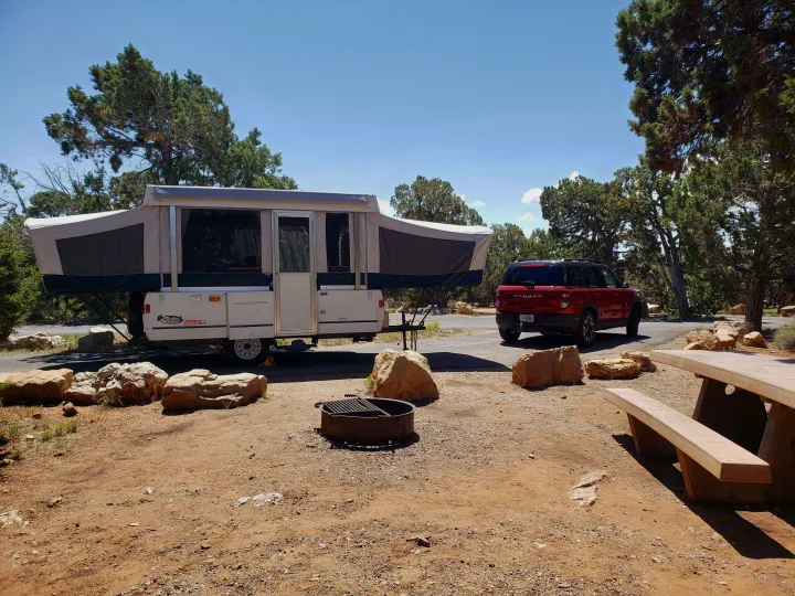 RV rental pop-up camper & SUV at a sunny campsite with a fire pit and concrete picnic table.. Coleman Sedona 1999