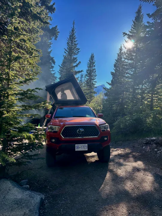 Red truck with rooftop tent set for forest camping. Person in tent. Enjoy nature, like an RV rental adventure, with mountain views & smoke.. Toyota Tacoma TRD OFF ROAD 2018