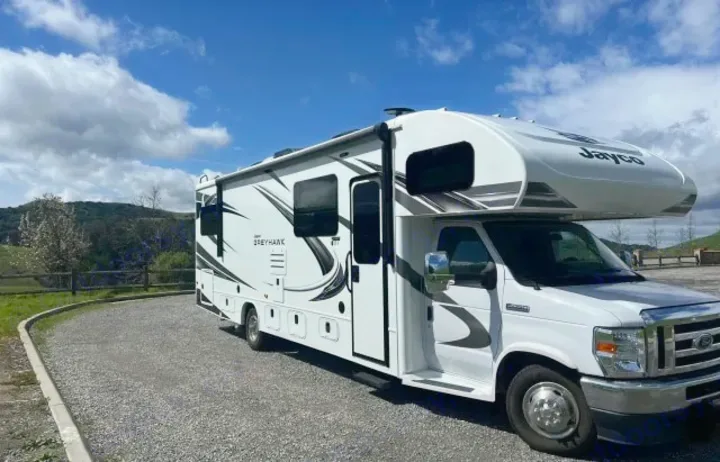 Modern white Jayco motorhome rv rental parked on gravel, with green hills and a cloudy blue sky.. Jayco Greyhawk 2021