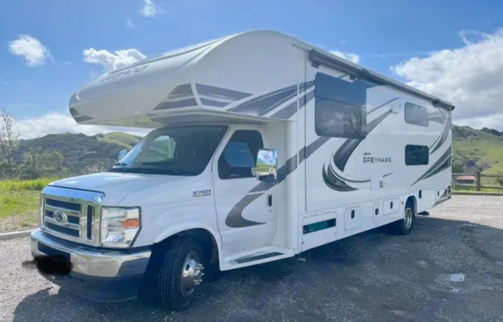 A white Jayco Greyhawk Class C RV rental is parked on a gravel lot with green hills and blue sky.. Jayco Greyhawk 2021