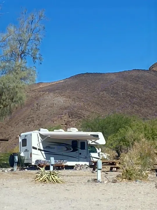 White RV rental with awning extended at a sunny desert campsite. Burnt mountains, sparse trees, and hookups are visible.. Forest River Sunseeker 2014