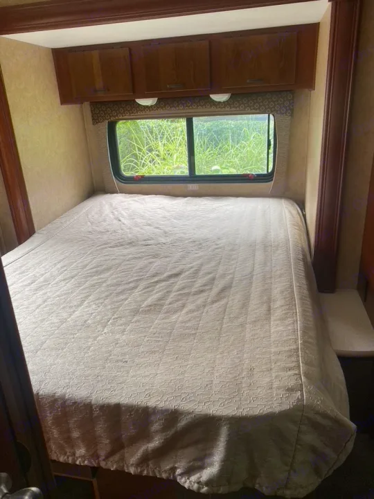 Cozy RV rental bedroom featuring a large bed with patterned comforter, window viewing nature, and overhead wood cabinets.. Forest River Sunseeker 2014