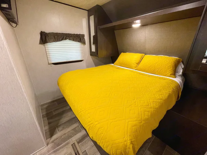 Bright and modern RV rental bedroom featuring a bed with a yellow quilted comforter, dark wood cabinetry, and a cozy window.. Heartland RVs Trail Runner SLE 2019