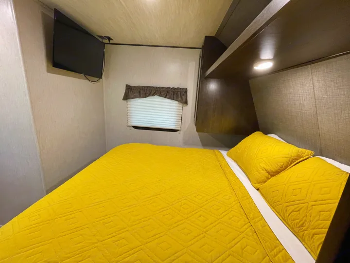 Cozy RV rental bedroom with a bright yellow quilted bed, wall-mounted TV, and overhead storage, perfect for your next trip.. Heartland RVs Trail Runner SLE 2019