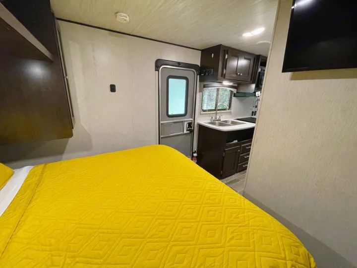 Inside an RV rental: bright yellow bed, compact kitchen with sink and window, and main entry door in a cozy interior.. Heartland RVs Trail Runner SLE 2019