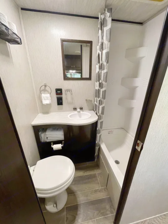 Compact RV rental bathroom featuring toilet, sink, mirror, and shower/tub combo with geometric curtain.. Heartland RVs Trail Runner SLE 2019