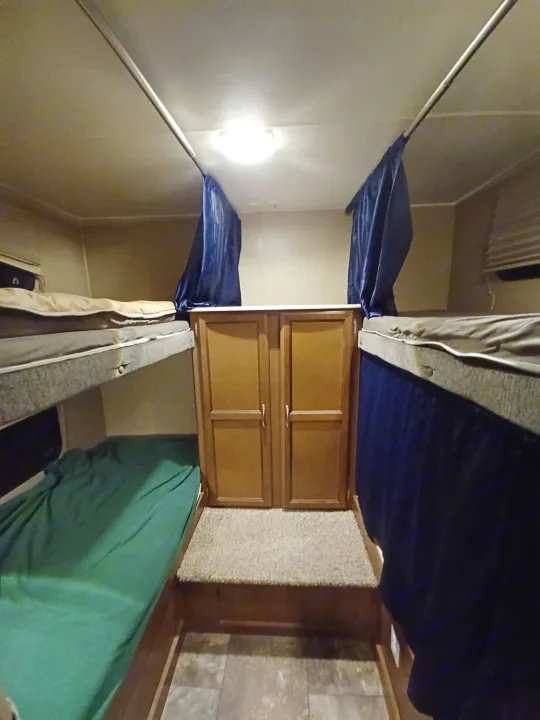 RV rental interior shows two sets of bunk beds with blue privacy curtains, flanking wood cabinets over carpet & tile.. Coachmen Freedom Express Liberty Edition 2016