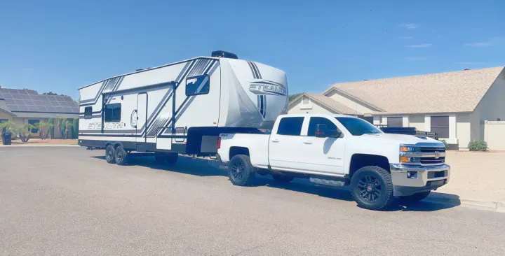 A powerful white pickup truck towing a large black and white Stealth fifth-wheel toy hauler on a sunny street. Ideal for your next rv rental.. Forest River Stealth 2020