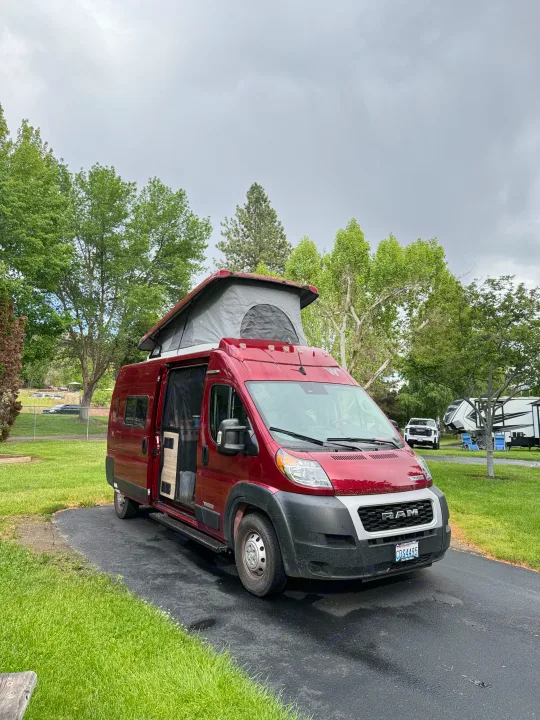 Red pop-top camper van (RV rental) parked on a wet driveway at a green campsite under cloudy skies.. Winnebago Solis 2023