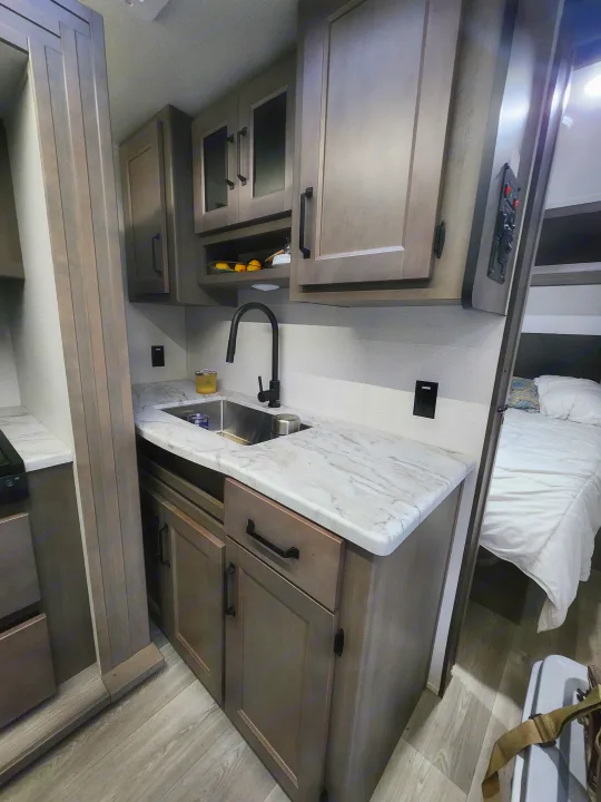 Modern RV rental interior featuring a compact kitchen with dark cabinets, a sink, and light counters, opening to a cozy bedroom.. Grand Design Transcend Xplor 2024