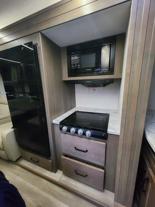 Compact kitchen in an RV rental, equipped with a Furrion microwave, stovetop, and refrigerator for easy travel cooking.. Grand Design Transcend Xplor 2024