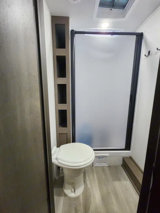 Compact RV rental bathroom with white toilet, retractable shower door, and built-in shelving, featuring light wood-look flooring.. Grand Design Transcend Xplor 2024
