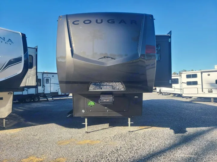 A dark Cougar fifth-wheel RV stands prominently on a sunny gravel lot, surrounded by other RVs, typical for an RV rental or dealership.. Keystone RV Cougar 2024