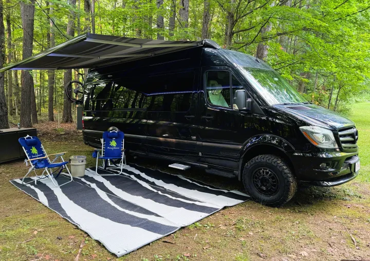 Black camper van with awning extended, chairs, and mat at a serene wooded campsite, perfect for an RV rental.. Airstream Interstate 2018
