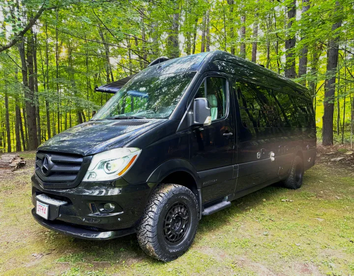 Black Airstream Sprinter RV rental, with off-road tires, parked in a vibrant, sunlit forest. Perfect for nature adventures.. Airstream Interstate 2018