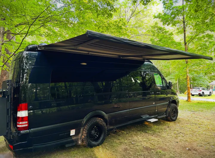 Black camper van with extended awning parked in a sunny forested campsite. Perfect for your next RV rental adventure.. Airstream Interstate 2018