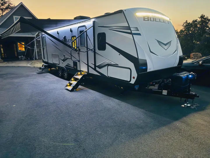 A modern Bullet RV rental, brightly lit with an extended awning and illuminated steps, ready for your next journey at dusk.. Keystone RV Bullet Ultra Lite 2023