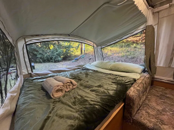 RV rental pop-up camper interior: a comfy bed with green sheets, folded towels, and a wide window showing a lush forest view.. Fleetwood Santa Fe 2004