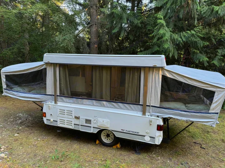 Pop-up RV rental ready for camping. White camper with extended beds extended, nestled in a lush, green outdoor setting.. Fleetwood Santa Fe 2004